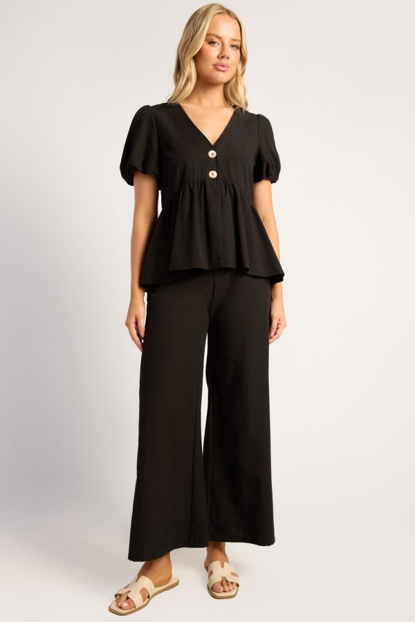 Model wearing the KoKo Romina black peplum top with puff sleeves, V-neckline and two contrast buttons, paired with matching wide-leg pants.